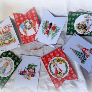 Peut inclure: Une guirlande de Noël avec des illustrations de lapins, de sapins et de scènes hivernales. Les fanions rouges et verts sont décorés de flocons de neige. Chaque fanion présente une illustration différente, reliée par une ficelle.