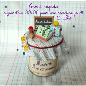 Peut inclure: Un bocal en verre avec une étiquette Merci Maîtresse, surmonté d'un couvercle en tissu à carreaux. Le dessus est décoré d'un crayon miniature, d'un petit livre, d'un cadeau avec un pompon jaune et d'une ardoise indiquant Année scolaire 2025-2026.