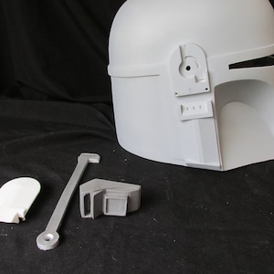 May include: A white Mandalorian helmet with separate components, including a visor, side pieces, and a connecting bar. The helmet has a smooth, matte finish and is designed for cosplay or display.