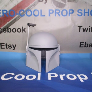 May include: A gray Mandalorian helmet prop with a T-shaped visor and a rangefinder. The helmet is displayed on a blue surface with the text "ZERO-COOL PROP SHOP" and social media logos in the background. The helmet is a replica.