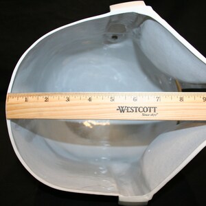 May include: A white, curved object with a gray interior is measured with a wooden ruler. The ruler shows a measurement of approximately 10 inches across the widest part of the object. A small, yellow foam block is in the background.