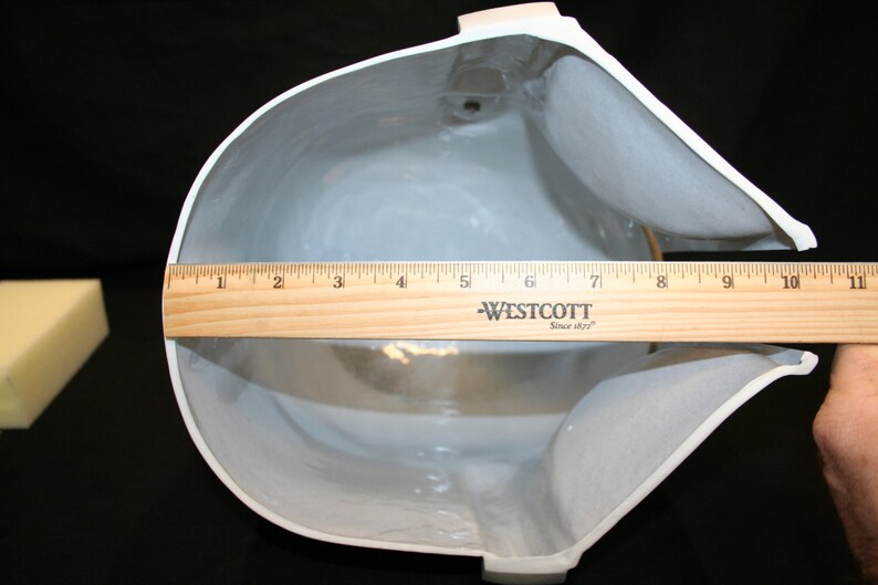 May include: A white, curved object with a gray interior is measured with a wooden ruler. The ruler shows a measurement of approximately 10 inches across the widest part of the object. A small, yellow foam block is in the background.