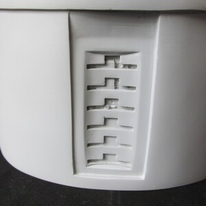 May include: Close-up of a light gray, cylindrical object with a recessed rectangular panel. The panel features a series of horizontal, stepped designs. The object has a smooth, matte finish and appears to be made of a solid material.