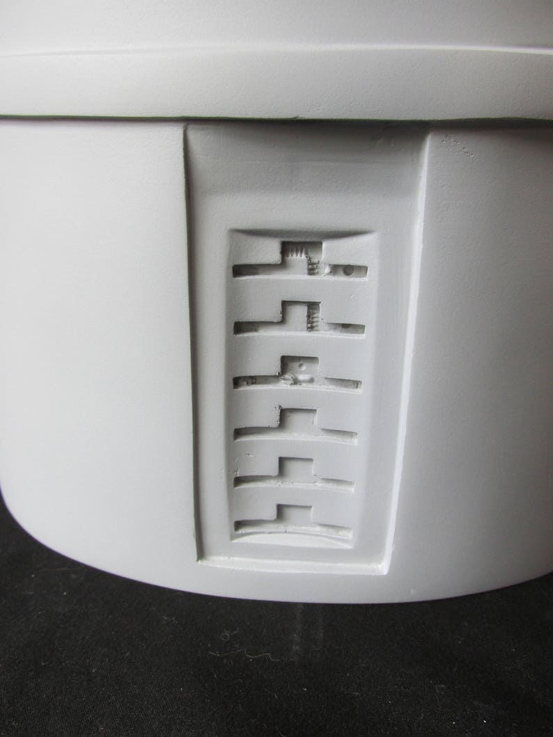 May include: Close-up of a light gray, cylindrical object with a recessed rectangular panel. The panel features a series of horizontal, stepped designs. The object has a smooth, matte finish and appears to be made of a solid material.
