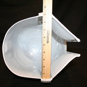 May include: A white, helmet-shaped object with a curved design, approximately 9 inches tall, is measured with a wooden ruler. The interior is visible, and a small, rectangular foam block is in the background. The object is on a black surface.