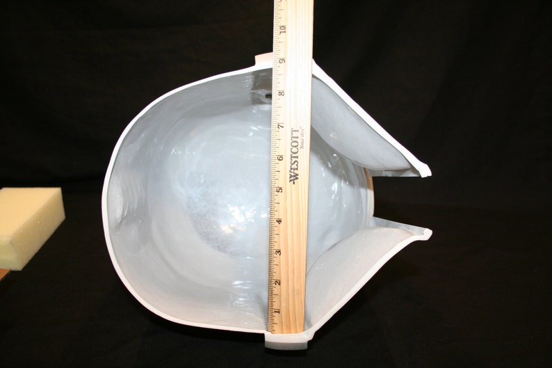 May include: A white, helmet-shaped object with a curved design, approximately 9 inches tall, is measured with a wooden ruler. The interior is visible, and a small, rectangular foam block is in the background. The object is on a black surface.