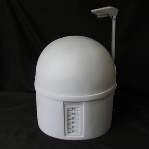 May include: A light gray helmet with a rounded top and a rectangular lower section. A vertical bar extends from the top with a small, flat rectangular piece at the end. The helmet has a series of rectangular indentations on the side.