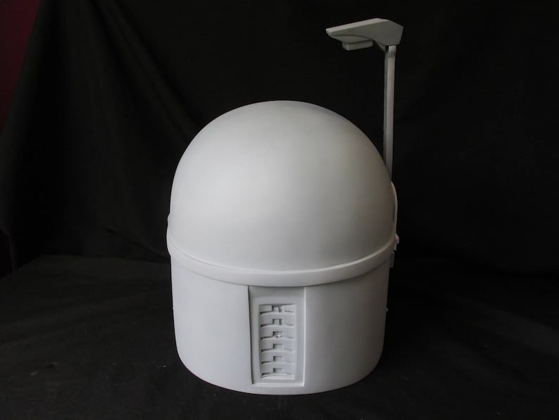 May include: A light gray helmet with a rounded top and a rectangular lower section. A vertical bar extends from the top with a small, flat rectangular piece at the end. The helmet has a series of rectangular indentations on the side.