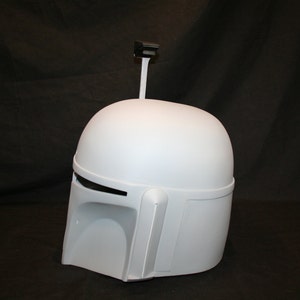May include: A light gray Mandalorian helmet with a rounded top, visor, and side details. A black rectangular antenna extends from the top. The helmet is a prop or costume piece.