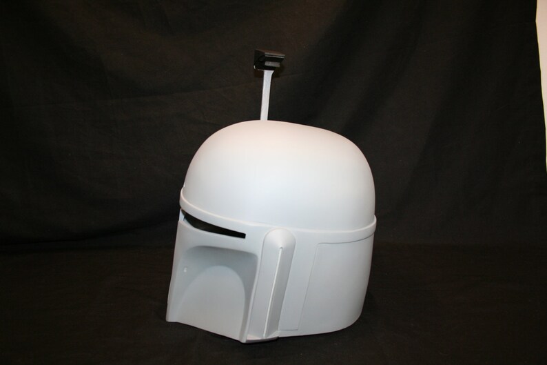 May include: A light gray Mandalorian helmet with a rounded top, visor, and side details. A black rectangular antenna extends from the top. The helmet is a prop or costume piece.