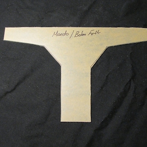 May include: A tan paper template in a T-shape, with the handwritten text "Mando / Boba Fett" across the top. The template is set against a black background.
