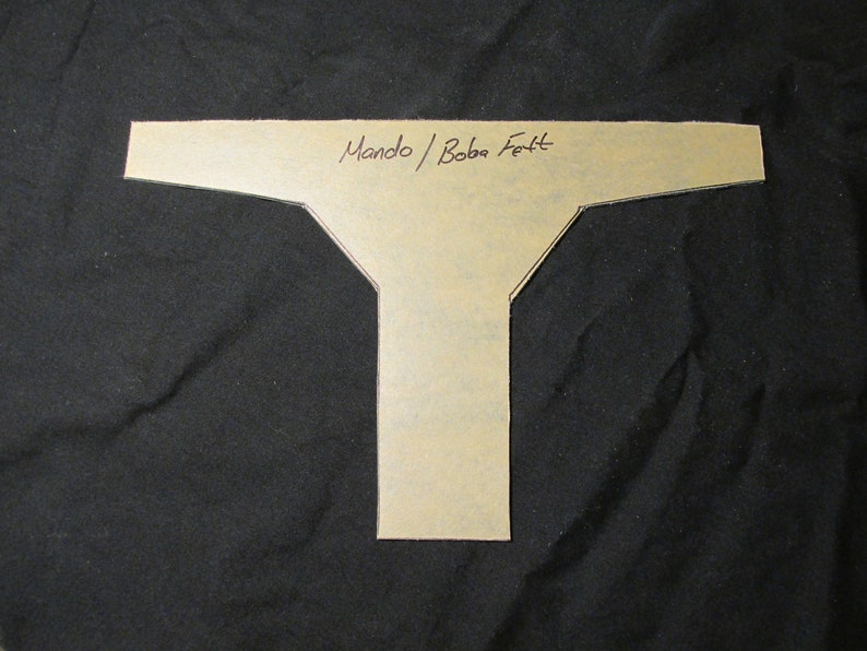 May include: A tan paper template in a T-shape, with the handwritten text "Mando / Boba Fett" across the top. The template is set against a black background.