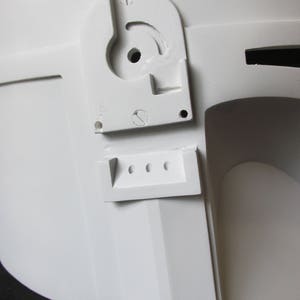 May include: Close-up of a white helmet with a detailed, angular design. Features include a curved, slotted component with a central hole and a rectangular piece with three circular openings. The helmet's surface is smooth and matte.