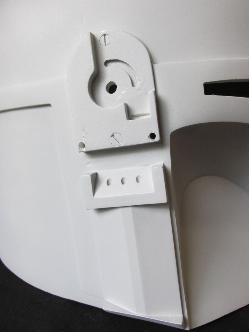 May include: Close-up of a white helmet with a detailed, angular design. Features include a curved, slotted component with a central hole and a rectangular piece with three circular openings. The helmet's surface is smooth and matte.