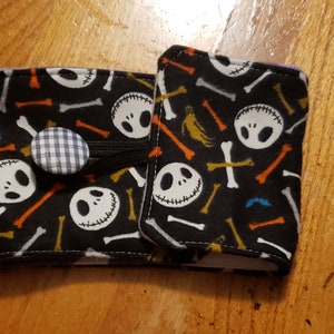 May include: A black and white fabric coffee cup cosy with a repeating pattern of Jack Skellington's face and bones. The cosy has a blue and white gingham button closure.