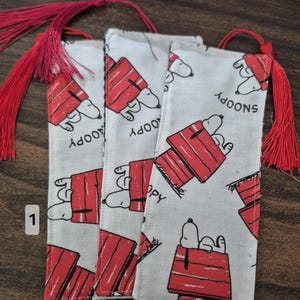 Peut inclure: Trois marque-pages rectangulaires en tissu avec un motif répétitif du personnage de dessin animé Snoopy allongé sur une niche rouge. Chaque marque-page est surmonté d'un pompon rouge vif attaché par un cordon rouge. Le tissu est blanc et noir.