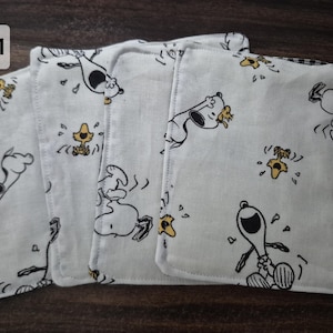 May include: A set of four square white fabric coasters featuring a repeating black and white line art pattern of the cartoon character Snoopy and the yellow bird Woodstock, with stitched edges and a soft, lightweight textile construction.