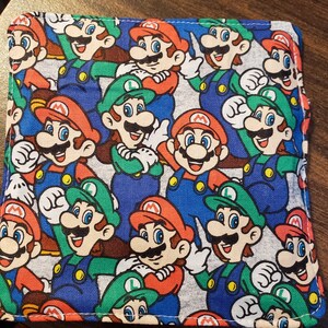 Pu&ograve; includere: Articolo in tessuto quadrato con un motivo ripetuto dei personaggi di Mario e Luigi nei loro abiti iconici. Il design include colori rosso, verde, blu e bianco. L'articolo sembra essere un portafoglio o un piccolo accessorio simile.