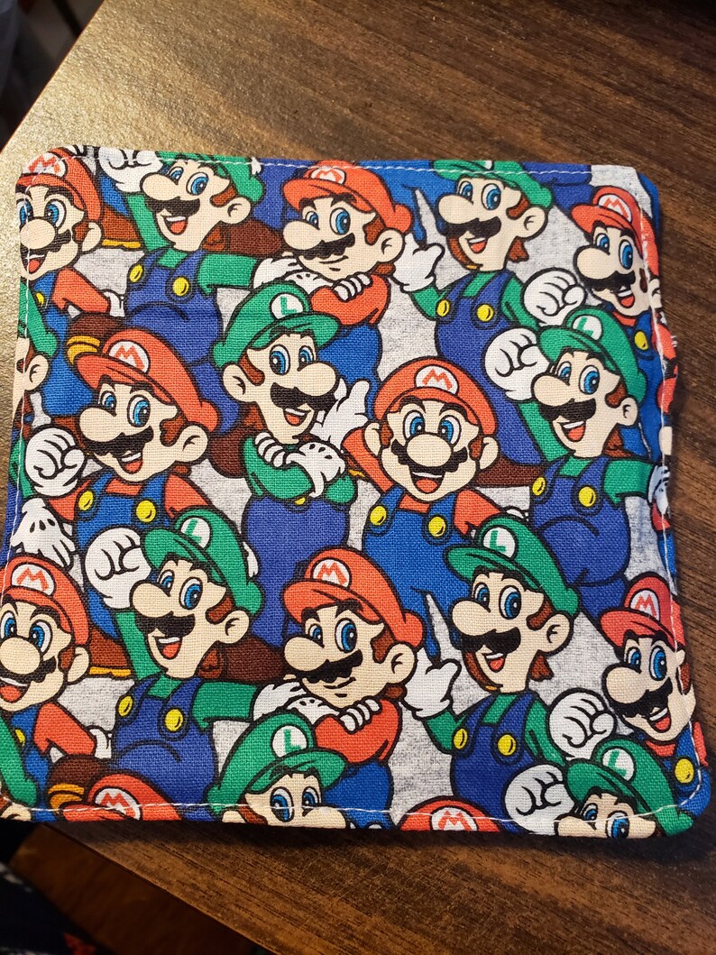 Pu&ograve; includere: Articolo in tessuto quadrato con un motivo ripetuto dei personaggi di Mario e Luigi nei loro abiti iconici. Il design include colori rosso, verde, blu e bianco. L'articolo sembra essere un portafoglio o un piccolo accessorio simile.
