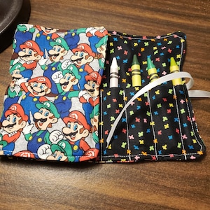 May include: A black fabric crayon holder with a colorful pattern and a white ribbon. The holder has four slots for crayons. The holder is open and shows four crayons: one white, one yellow, and two green.