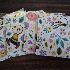 Pu&ograve; includere: Quattro tovaglioli di stoffa bianchi con disegni floreali e di personaggi dei cartoni animati. I tovaglioli presentano Snoopy, Charlie Brown e altri personaggi dei Peanuts. Il motivo floreale comprende fiori rosa, viola e gialli con foglie verdi.