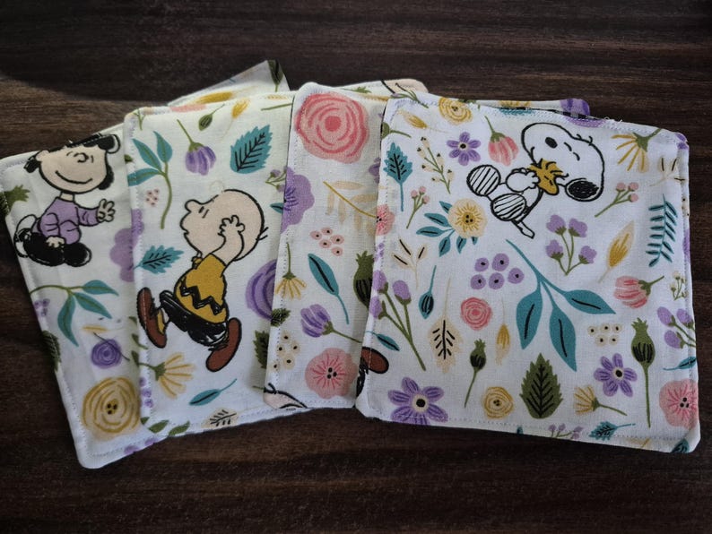 Pu&ograve; includere: Quattro tovaglioli di stoffa bianchi con disegni floreali e di personaggi dei cartoni animati. I tovaglioli presentano Snoopy, Charlie Brown e altri personaggi dei Peanuts. Il motivo floreale comprende fiori rosa, viola e gialli con foglie verdi.