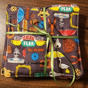 Pu&ograve; includere: Un set di sottobicchieri quadrati in tessuto con sfondo grigio scuro e un motivo colorato di ombrelli, aragoste e il logo Central Perk. I sottobicchieri sono legati con un nastro verde.