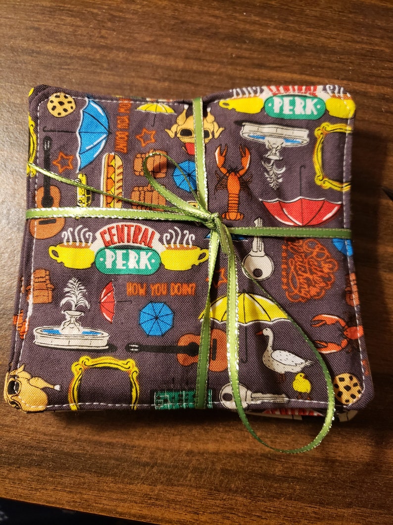 Pu&ograve; includere: Un set di sottobicchieri quadrati in tessuto con sfondo grigio scuro e un motivo colorato di ombrelli, aragoste e il logo Central Perk. I sottobicchieri sono legati con un nastro verde.