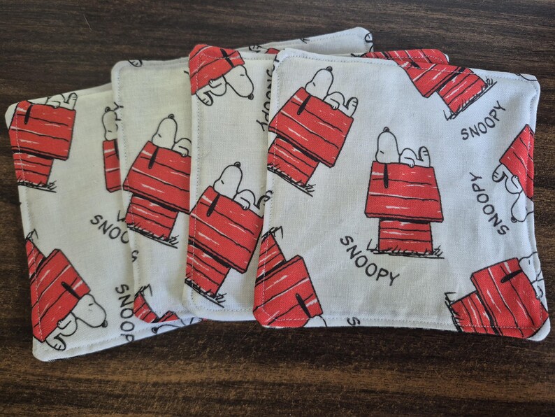 Pu&ograve; includere: Set di sottobicchieri quadrati in tessuto con un motivo ripetuto di Snoopy nella sua cuccia rossa. La parola "SNOOPY" &egrave; stampata sui sottobicchieri. I sottobicchieri sono bianchi con dettagli rossi e neri.