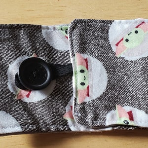 May include: A fabric coffee cup sleeve with a black button fastening. The sleeve has a repeating pattern of white circles with green and pink cartoon characters on a grey denim-like background.