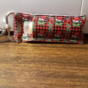 May include: A red and black plaid fabric lip balm holder with a Grinch design. The holder has a silver metal clip.