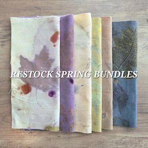 May include: Six fabric bundles in shades of cream, purple, and blue, each with leaf imprints. The image displays the text "RESTOCK SPRING BUNDLES". The fabrics are arranged vertically on a wooden surface.