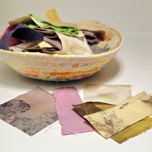 May include: A white woven basket filled with scraps of fabric in various colors and patterns. The basket is sitting on a white surface, and there are several smaller pieces of fabric in different colors and patterns scattered around it.