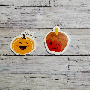 Pode incluir: Dois adesivos de desenho animado, um de uma abóbora sorridente e outro de uma maçã caramelada piscando. Ambos os adesivos têm contornos brancos e estão sobre um fundo de madeira.