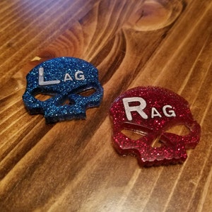 May include: Two glitter skull dice with the letters "LAG" and "RAG" on them. The skull on the left is blue and the skull on the right is red.