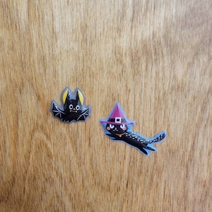 May include: Two black Halloween-themed stickers on a wood background. One sticker is a bat with yellow ears and the other is a black cat wearing a witch's hat.