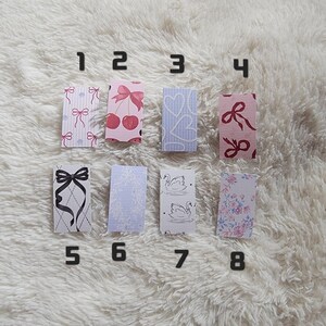 Op de afbeelding: Acht rechthoekige decoratieve stickers met verschillende ontwerpen, waaronder strikken, kersen, harten en bloempatronen. De stickers zijn genummerd van 1 tot 8 en zijn gerangschikt op een zacht, wit oppervlak.