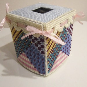 May include: A square tissue box cover with a patchwork design. The cover is made of white fabric with a variety of colors including pink, blue, yellow, green, and purple. The cover has a white lace trim and pink ribbons.