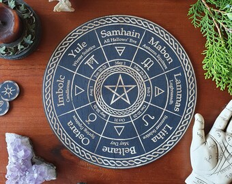 May include: A wooden wheel with a black background and gold lettering. The wheel is divided into 12 sections, each with a different name for a pagan holiday. The names of the holidays are: Yule, Winter Solstice, Samhain, All Hallows' Eve, Mabon, Autumn Equinox, Lammas, Lughnasadh, Litha, Summer Solstice, Beltane, Ostara, Imbolc, and Candlemas.