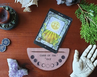 Könnte beinhalten: Ein hölzerner Kartenhalter für den Tag mit einem Halbmond-Design und dem Text "Card of the Day". Der Halter steht auf einer Holzoberfläche mit einer Tarotkarte, die eine Sonne mit einem Gesicht und den Text "The Sun" zeigt.