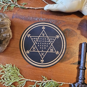 Könnte beinhalten: Ein schwarzer Holzkreis mit einem goldenen Stern und dem Wort "ABRACADABRA" in einem Kreis darum herum geschrieben. Der Kreis ist von einem schwarzen Rand und einem goldenen Außenring umgeben.