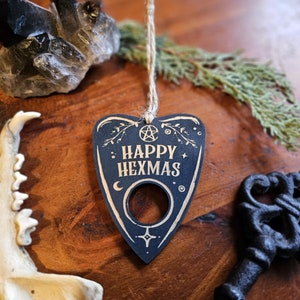 May include: Black wooden ornament shaped like a witch's board with the text "HAPPY HEXMAS" in white. The ornament has a hole in the center and is hanging from a twine string.