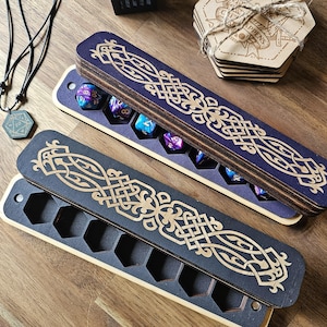 Peut inclure: Une boîte en bois avec un motif de nœud celtique bleu et or. La boîte a un couvercle à charnière et est ouverte pour révéler un jeu de sept dés. Les dés sont violets et bleus avec des paillettes dorées.