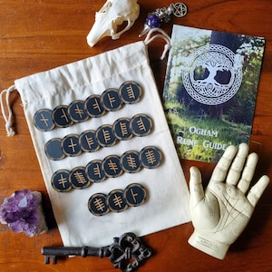 May include: A set of 15 black and gold Ogham runes, with a white cloth bag, a book titled "Ogham Rune Guide", and a palmistry hand. The runes are circular and feature a different symbol on each one.