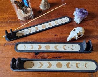 Puede incluir: Tres porta inciensos de madera negra con un diseño de fases lunares blancas. Los porta inciensos están dispuestos en fila sobre una superficie de madera.