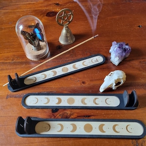 Könnte beinhalten: Drei schwarze Holz-Räucherstäbchenhalter mit einem weißen Mondphasen-Design. Die Halter sind in einer Reihe auf einer Holzoberfläche angeordnet.