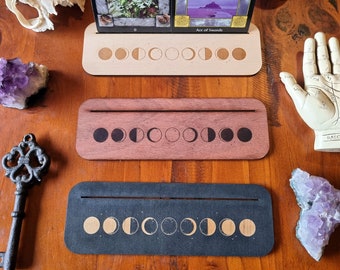 May include: Three wooden tarot card holders with moon phase designs. The top holder is light-colored, the middle is reddish-brown, and the bottom is dark gray. Tarot cards are displayed in the top holder. Additional spiritual items are in the background.