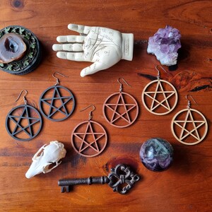 May include: A set of six wooden earrings featuring a pentacle design. The earrings are in two different colors: brown and black. The earrings are displayed on a wooden surface with other decorative items.