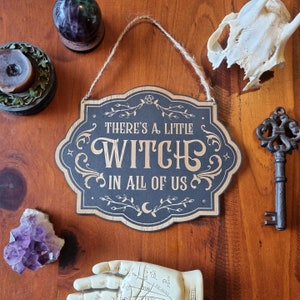 Op de afbeelding: Een zwart houten bord met gouden letters met de tekst "There's a little witch in all of us." Het bord heeft een decoratieve rand met wijnstokken en een halve maan.