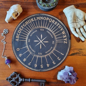 May include: A black and gold Ouija board with the letters of the alphabet, numbers 0-9, and the words "yes", "no", "rephrase", and "unknown" printed on it. The board is surrounded by various objects, including a crystal, a key, and a hand.